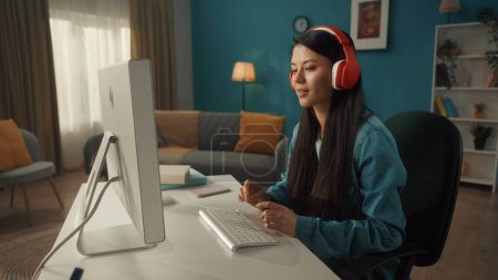 Téléchargez les photos : Vue latérale d'une femme asiatique en casque sans fil rouge travaillant sur un ordinateur. Une jeune femme joue à un jeu en ligne, travaille, regarde des vidéos, écoute de la musique. Gros plan - en image libre de droit