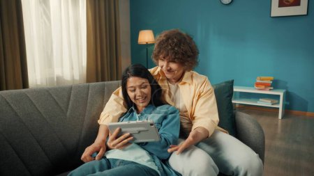 Photo for A young married couple spends time together on a day off. a Woman and a man watch a movie, photo, video on a tablet. A young Asian woman lies on a sofa with a tablet in her hands, leaning her back on - Royalty Free Image