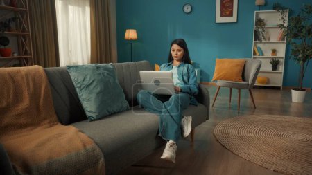 Foto de Atractiva mujer asiática está sentada en el sofá de la sala de estar con un ordenador portátil en su regazo. Una mujer trabaja, navega por Internet, mira fotos, videos. Trabajo remoto, freelancer - Imagen libre de derechos