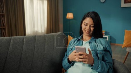 Photo for A young Asian woman sits on a sofa with a phone in her hands. A woman is typing a message, chatting, browsing social networks, scrolling through the feed. Portrait of a smiling woman in the living - Royalty Free Image
