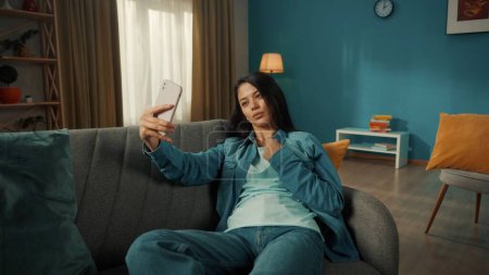 Téléchargez les photos : Attrayant asiatique femme prend un selfie sur un smartphone, pose. Une jeune femme est assise sur un canapé dans le salon avec un téléphone dans les mains - en image libre de droit