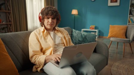 Téléchargez les photos : Un jeune homme est assis sur un canapé portant des écouteurs sans fil rouges avec un ordinateur portable sur les genoux. Un homme tape un texte, un message, bavarde, écoute de la musique, joue à des jeux en ligne. Gros plan - en image libre de droit