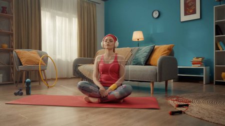 Foto de Una joven y atractiva mujer se sienta con las piernas cruzadas en posición de loto con los ojos cerrados. Una mujer pelirroja con un chándal rosa, diadema y auriculares inalámbricos blancos practica yoga en casa. Deportes - Imagen libre de derechos