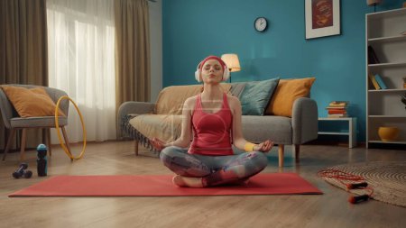 Téléchargez les photos : Une jeune femme séduisante s'assoit les jambes croisées en position lotus les yeux fermés. Une femme rousse en survêtement rose, bandeau et casque sans fil blanc pratique le yoga à la maison. Sports - en image libre de droit