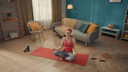 Téléchargez les photos : Une jeune femme s'assoit les jambes croisées en position lotus les yeux fermés. Une femme rousse en survêtement rose, bandeau et casque sans fil blanc pratique le yoga à la maison. Vue d'en haut. Sports - en image libre de droit