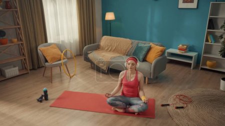 Téléchargez les photos : Une jeune femme s'assoit les jambes croisées en position lotus les yeux fermés. Une femme rousse en survêtement rose, bandeau et casque sans fil blanc pratique le yoga à la maison. Vue d'en haut. Sports - en image libre de droit