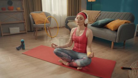 Foto de Una joven y atractiva mujer se sienta con las piernas cruzadas en posición de loto con los ojos cerrados. Una mujer pelirroja con un chándal rosa, diadema y auriculares inalámbricos blancos practica yoga en casa. Deportes - Imagen libre de derechos