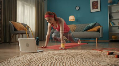 Eine Frau treibt Sport, Bergsteigerin, rennt im Liegen, auf dem Boden vor einem Laptop-Bildschirm. Die junge Frau beugte ein Bein an den Knie- und Hüftgelenken, das andere entkrümmte und