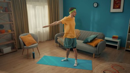 Photo for Sports at home. A man in sportswear and with a green headband is warming up in the living room. The man took his straight arms back, connecting the shoulder blades and making turns of the torso. Home - Royalty Free Image