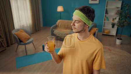 Photo for Profile portrait of a man in a tracksuit and yellow sports headband in a living room with sports equipment on the floor. The man holds a glass of juice in his hand, fixing his eyes on it. Close up - Royalty Free Image