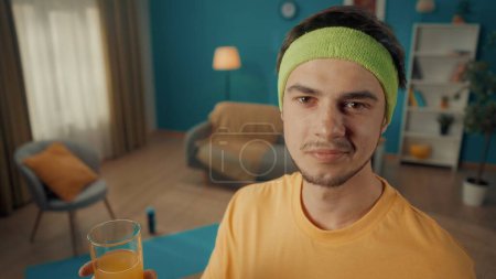Photo for Portrait of a man in a tracksuit and yellow sports headband in a living room with sports equipment on the floor. The man holds a glass of juice in his hand and looks straight ahead. Close up - Royalty Free Image