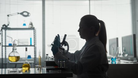 Foto de Laboratorio científico médico. Una investigadora aplica una muestra a una diapositiva de vidrio, una micropreparación usando una pipeta. Silueta oscura de una científica cerca de un microscopio en un laboratorio - Imagen libre de derechos