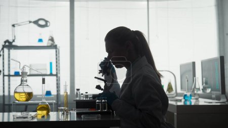 Foto de Laboratorio científico médico. Vista lateral de una silueta oscura de una científica mirando bajo un microscopio, haciendo un análisis de una muestra de prueba. Especialista en biotecnología ambicioso, trabajando con - Imagen libre de derechos
