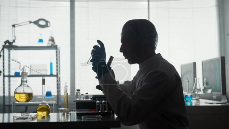Foto de Laboratorio científico médico. Un hombre investigador aplica una muestra a una diapositiva de vidrio, una micropreparación usando una pipeta. Silueta oscura de un científico masculino cerca de un microscopio en un laboratorio - Imagen libre de derechos