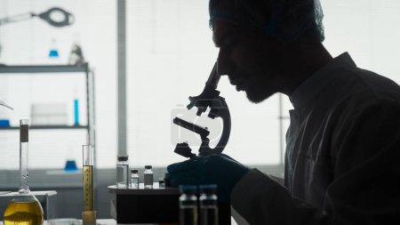 Foto de Laboratorio científico médico. Un joven científico en un laboratorio de investigación examina una muestra en un micropreparado usando un microscopio. Vista lateral de la silueta oscura de un hombre sentado frente a un - Imagen libre de derechos