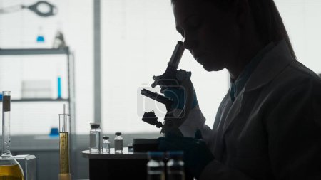 Foto de Laboratorio científico médico. Vista lateral de una silueta oscura de una científica mirando bajo un microscopio, haciendo un análisis de una muestra de prueba de cerca. Especialista en biotecnología ambiciosa, trabajando - Imagen libre de derechos