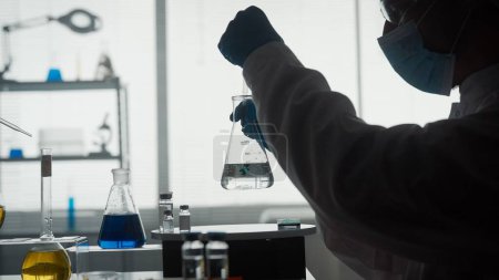 Foto de Silueta oscura de un científico masculino llevando a cabo un experimento científico de cerca. Un hombre usando una pipeta agregará un reactivo verde a un frasco de vidrio con un líquido transparente - Imagen libre de derechos