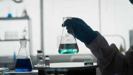 Foto de Experimento científico. Un frasco con un líquido transparente y un reactivo verde en una mano de hombre de cerca - Imagen libre de derechos