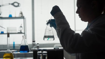 Foto de Silueta oscura de una científica que lleva a cabo un experimento científico de cerca. Una mujer que usa una pipeta agregará un reactivo verde a un frasco de vidrio con un líquido transparente - Imagen libre de derechos