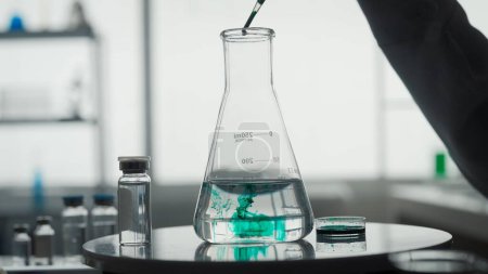 Foto de Experimento científico. Un frasco con un líquido transparente se coloca sobre una mesa sobre un fondo borroso del laboratorio de cerca. La mano de los científicos, usando una pipeta, añade un reactivo verde al frasco - Imagen libre de derechos