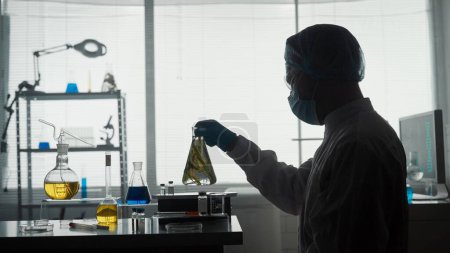Foto de Un científico masculino sostiene un frasco Erlenmeyer con una planta dentro y lo examina. Silueta oscura de un científico con un frasco en las manos en el laboratorio. Experimento científico - Imagen libre de derechos