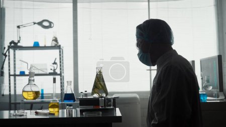 Foto de Un científico masculino examina un frasco Erlenmeyer con una planta dentro. Silueta oscura de un científico con una bata blanca, gorra, gafas y una máscara en el laboratorio. Experimento científico - Imagen libre de derechos
