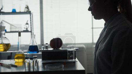 Foto de Silueta oscura de una científica sentada en una mesa frente a una manzana, una jeringa y una ampolla de un químico. La mujer está trabajando en modificaciones de frutas, enriquecimiento de sabores, resistencia a - Imagen libre de derechos