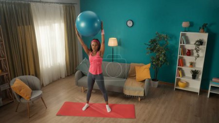 Photo for African American woman exercising with a fitness ball. An athletic woman raised a fitball over her head on straight outstretched arms. Young woman goes in for sports at home. Sport concept, home - Royalty Free Image