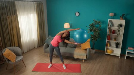 Photo for African American woman exercising with a fitness ball. A sporty woman does side bends with a fitball in her hands. Young woman goes in for sports at home. Sport concept, home fitness. View from above - Royalty Free Image