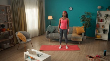 Photo for An African American woman in a tracksuit and headband stands on a sports mat in the living room. A young woman is looking at a laptop on a table. A woman watches a video tutorial, an online workout - Royalty Free Image