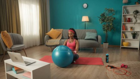Foto de Una mujer afroamericana en chándal y diadema se sienta con las piernas cruzadas en una alfombra deportiva en la sala de estar. Una mujer joven sostiene una pelota de fitness frente a ella y mira un portátil en la mesa. Mujer. - Imagen libre de derechos