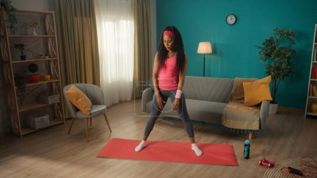 Téléchargez les photos : Une Afro-Américaine en survêtement et bandeau se tient sur un tapis de sport dans le salon. Une jeune femme se tient debout, les pieds écartés, prête à faire un exercice sportif. Le concept - en image libre de droit