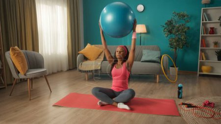 Photo for A young woman sits cross legged on a sports mat, on the floor in the living room. An African American woman raised her straight arms with a fitness ball above her head. Home fitness concept - Royalty Free Image