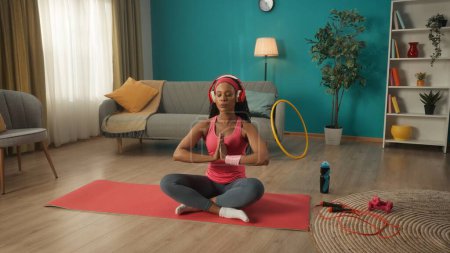 Téléchargez les photos : Une Afro-Américaine en casque rouge est assise sur un tapis de sport dans le salon. La femme s'assoit en position lotus, écoute de la musique, médite, se détend. Concept sportif. Cours de yoga - en image libre de droit