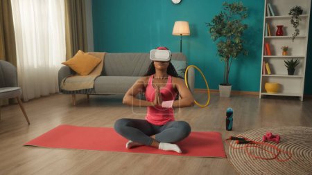 Photo for African American woman in virtual reality glasses sits in a lotus position. A young woman in a pink tracksuit is doing yoga in virtual reality. Lifestyle and technology concept - Royalty Free Image