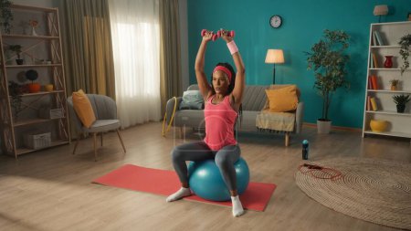 Foto de Mujer afroamericana sentada en el fitball y haciendo ejercicio con pesas en casa. Sentada en una pelota de fitness, una mujer entrena los músculos de sus brazos y hombros. Concepto deportivo, fitness en casa - Imagen libre de derechos