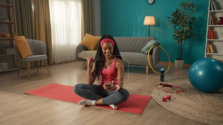 Téléchargez les photos : Une Afro-Américaine est assise les jambes croisées sur un tapis de sport par terre dans le salon. Une femme tient un haltère dans une main, exerçant sa main, et un smartphone dans l'autre main. Un sourire - en image libre de droit