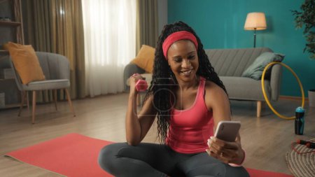 Foto de Mujer afroamericana sentada en una alfombra deportiva en la sala de estar de cerca. Una mujer sostiene una pesa en una mano, ejercitando su mano, y un teléfono inteligente en la otra mano. Una mujer sonriente está mirando - Imagen libre de derechos