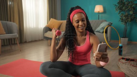 Téléchargez les photos : Femme afro-américaine assise sur un tapis de sport dans le salon de près. Une femme tient un haltère dans une main, exerçant sa main, et un smartphone dans l'autre main. Une femme souriante regarde - en image libre de droit