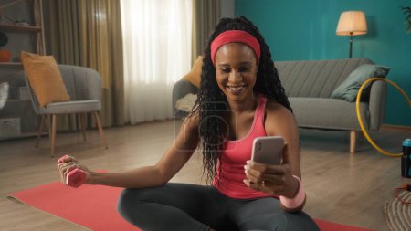 Téléchargez les photos : Femme afro-américaine assise sur un tapis de sport dans le salon de près. Une femme tient un haltère dans une main, exerçant sa main, et un smartphone dans l'autre main. Une femme souriante regarde - en image libre de droit