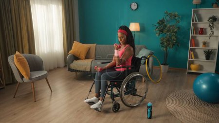 Foto de Mujer afroamericana en silla de ruedas haciendo ejercicio con pesas en la sala de estar. Una mujer con una diadema rosa y un chándal entrena los músculos de sus brazos, sacude sus bíceps y tríceps. El - Imagen libre de derechos