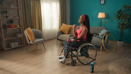 Photo for African American woman in a wheelchair exercising with dumbbells in the living room. A woman in a pink headband and in a tracksuit trains the muscles of her arms, shakes her biceps and triceps. The - Royalty Free Image