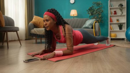 Photo for African American woman in sportswear lies on the floor in front of the phone. A woman performs an elbow plank exercise. Home fitness concept, online sports class - Royalty Free Image