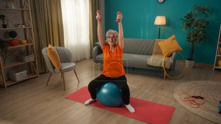 Foto de Una mujer jubilada de pelo gris sentada en una pelota de fitness realiza un ejercicio con pesas. Una anciana levanta los brazos rectos con pesas por encima de su cabeza. Manteniendo el cuerpo en tono. Concepto de - Imagen libre de derechos
