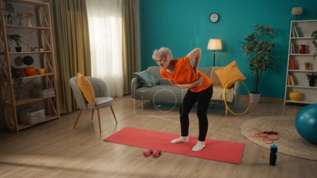 Photo for Chronic, agerelated, muscular back pain. An elderly woman is leaning over dumbbells and holding on to her back and side. Retired woman holding lower back in pain while standing in living room - Royalty Free Image
