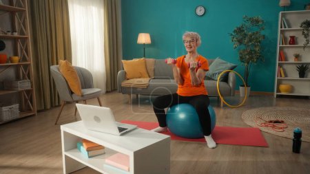 Photo for Elderly woman goes in for sports sitting on a fitness ball in front of a laptop. An elderly woman holds dumbbells in front of her in her arms, bent at the elbows. Home fitness concept, online sports - Royalty Free Image