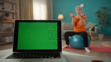Téléchargez les photos : Un ordinateur portable avec un écran vert sur une table sur un fond flou dans le salon fermer. Une femme âgée est assise sur une balle de fitness, faisant un exercice avec des haltères. Mode de vie, technologie - en image libre de droit