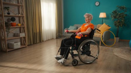 Téléchargez les photos : Femme âgée en fauteuil roulant faisant de l'exercice avec des haltères dans le salon. Une vieille femme aux cheveux gris entraîne les muscles des bras. Le concept de réadaptation physique - en image libre de droit