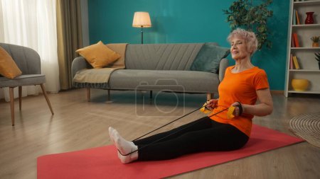 Téléchargez les photos : Femme aux cheveux gris faisant de l'exercice avec une bande élastique dans le salon. Une femme âgée s'assoit les jambes sur le sol. L'extenseur est enroulé derrière les pieds des femmes - en image libre de droit