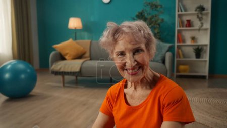 Téléchargez les photos : Portrait d'une femme âgée souriante contentante dans le salon de près. Une retraitée aux cheveux gris, fatiguée, mais satisfaite après une séance d'entraînement. Le concept de style de vie sportif, retraite, santé - en image libre de droit